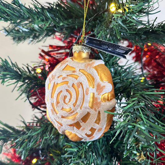 Glass Pastry Cinnamon Roll Ornament, a golden glass cinnamon roll with a white frosted swirl, hanging on a Christmas tree