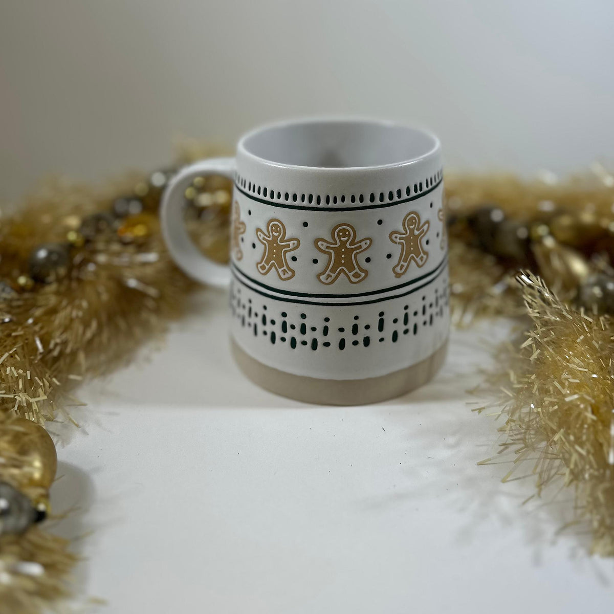 Gingerbread Man Mug, a white stoneware mug with gingerbread men and green dotted stripes, surrounded by gold tinsel and baubles