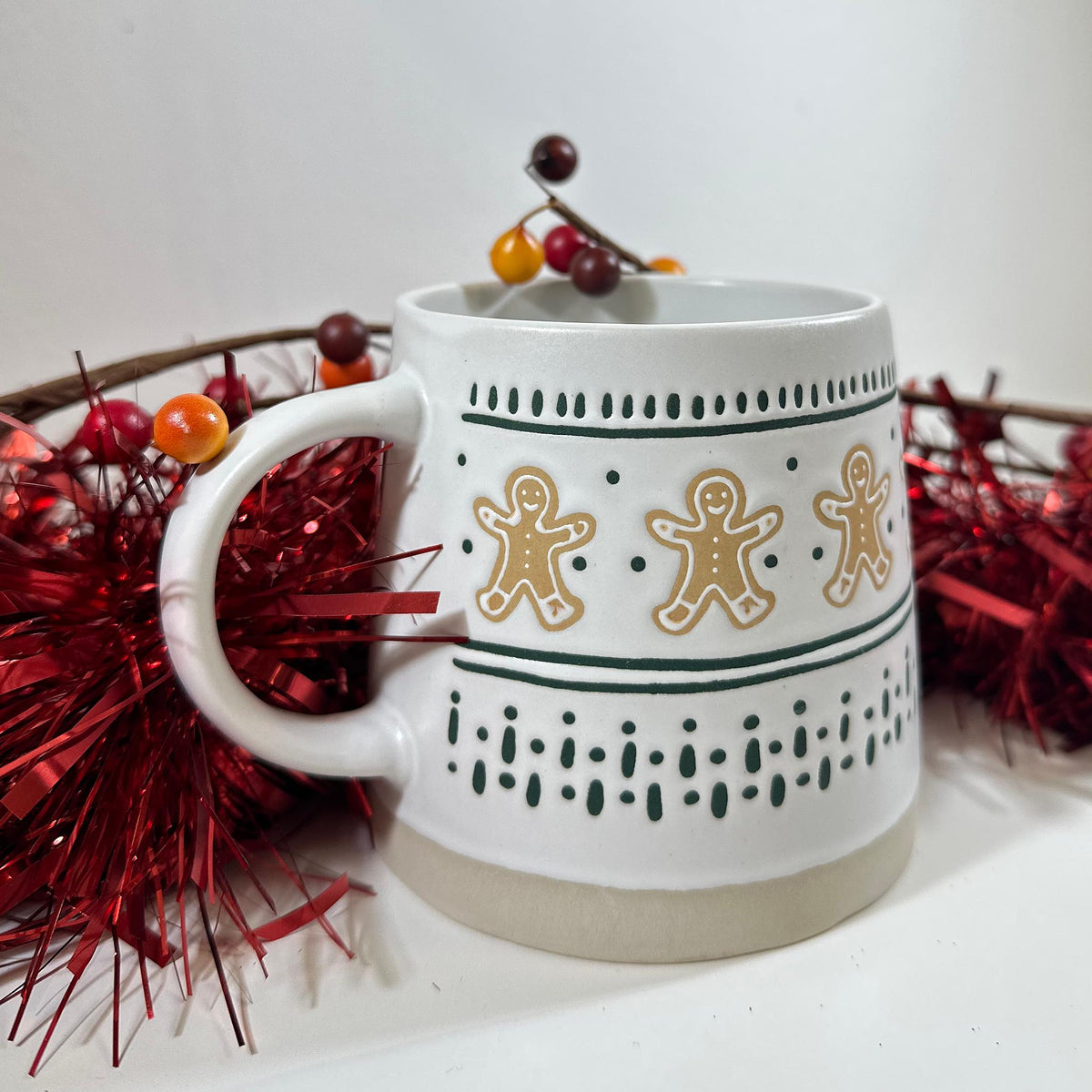 Gingerbread Man Mug, a white stoneware mug with gingerbread men and green dotted stripes, on red tinsel with a berry sprig