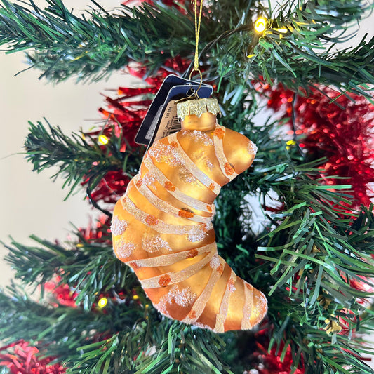 Glass Pastry Croissant Ornament, a golden glass croissant with white stripes and speckles, hanging on a Christmas tree