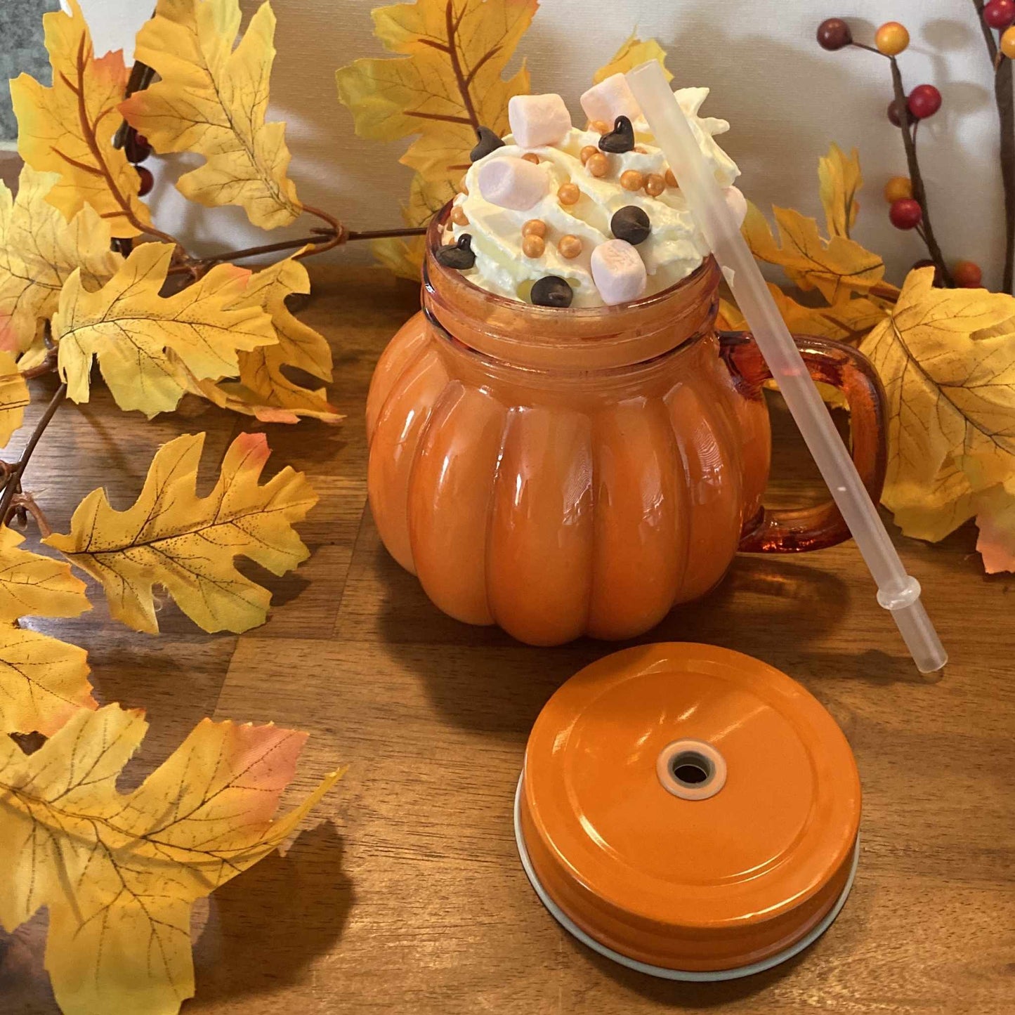 Orange pumpkin-shaped glass mug with lid and straw, topped with whipped cream and marshmallows, surrounded by autumn leaves